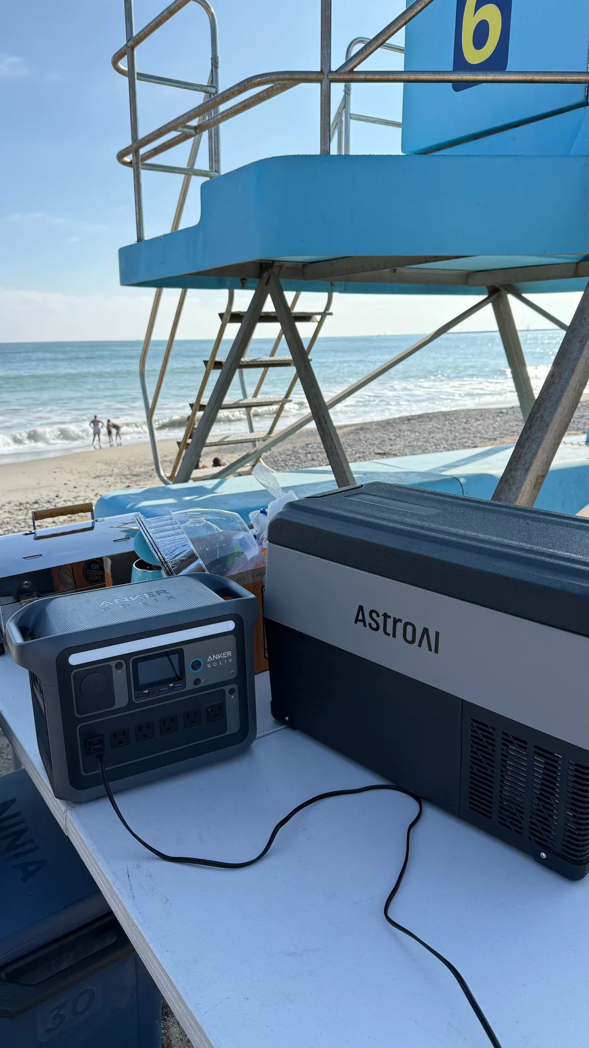 Anker portable power station and AstroAI portable refrigerator set up on a white folding table at the beach