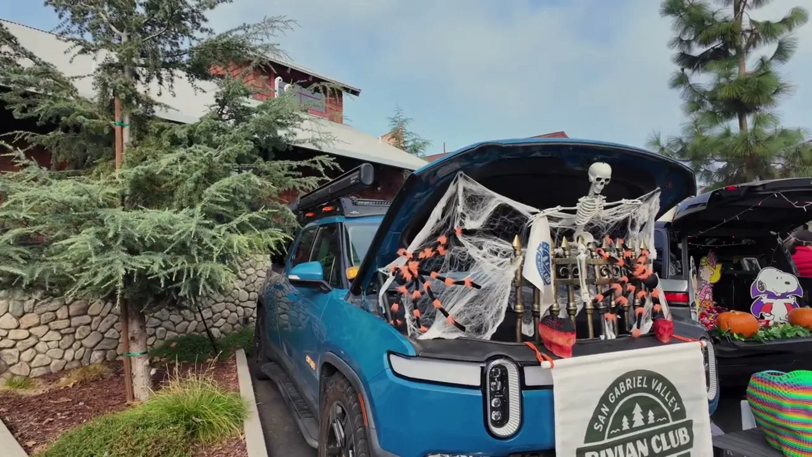 Blue Rivian R1T with a skeleton and spider web Halloween frunk display behind the San Gabriel Valley Rivian Club banner