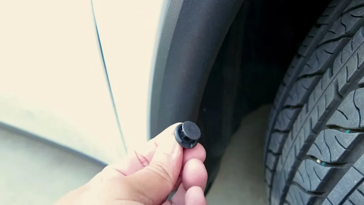 Hand holding a black plastic push-pin fastener near a white car fender and tire.