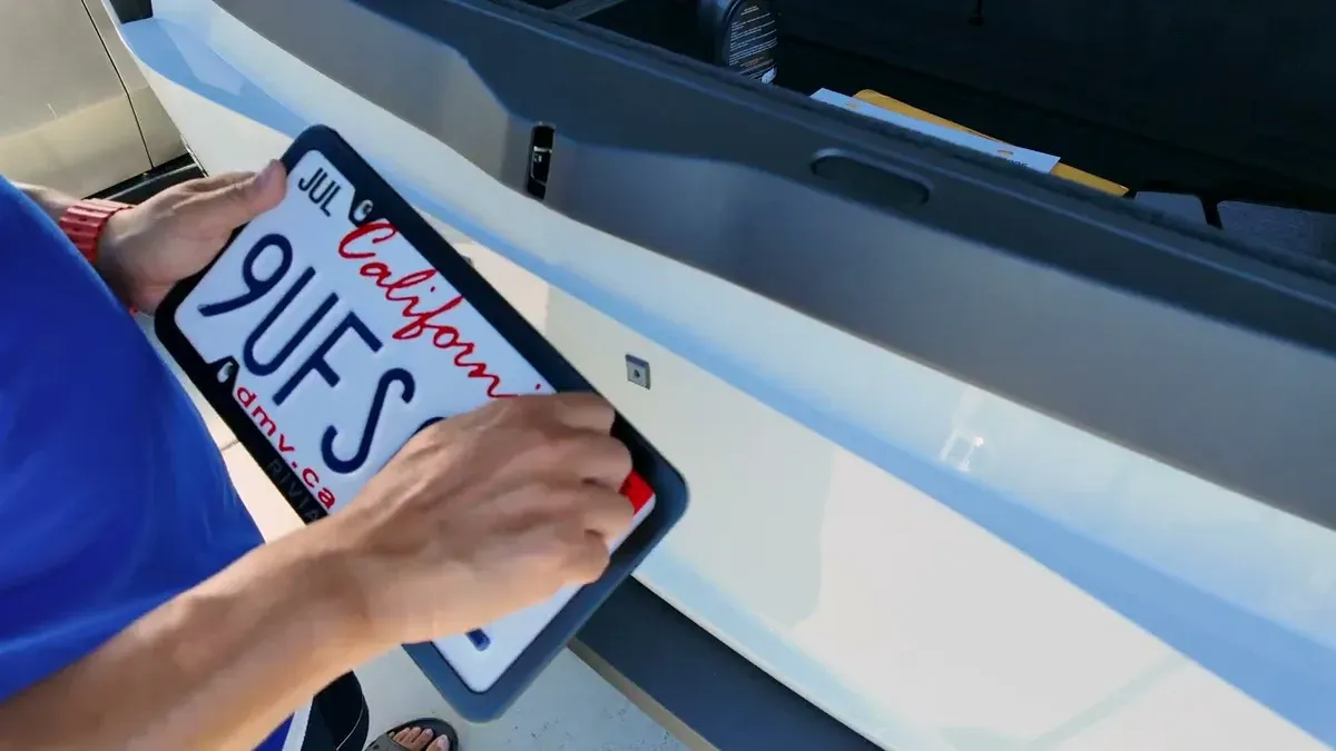 Close-up of hands securing a black-framed California license plate to the white Rivian R1S.
