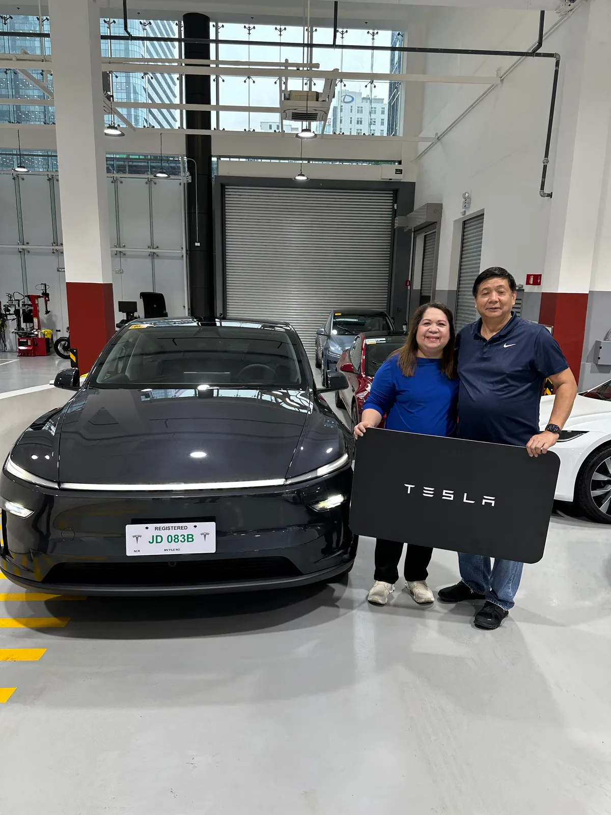 Doc Dennis and his wife holding a Tesla sign in front of their new Model Y at the Tesla delivery center in BGC