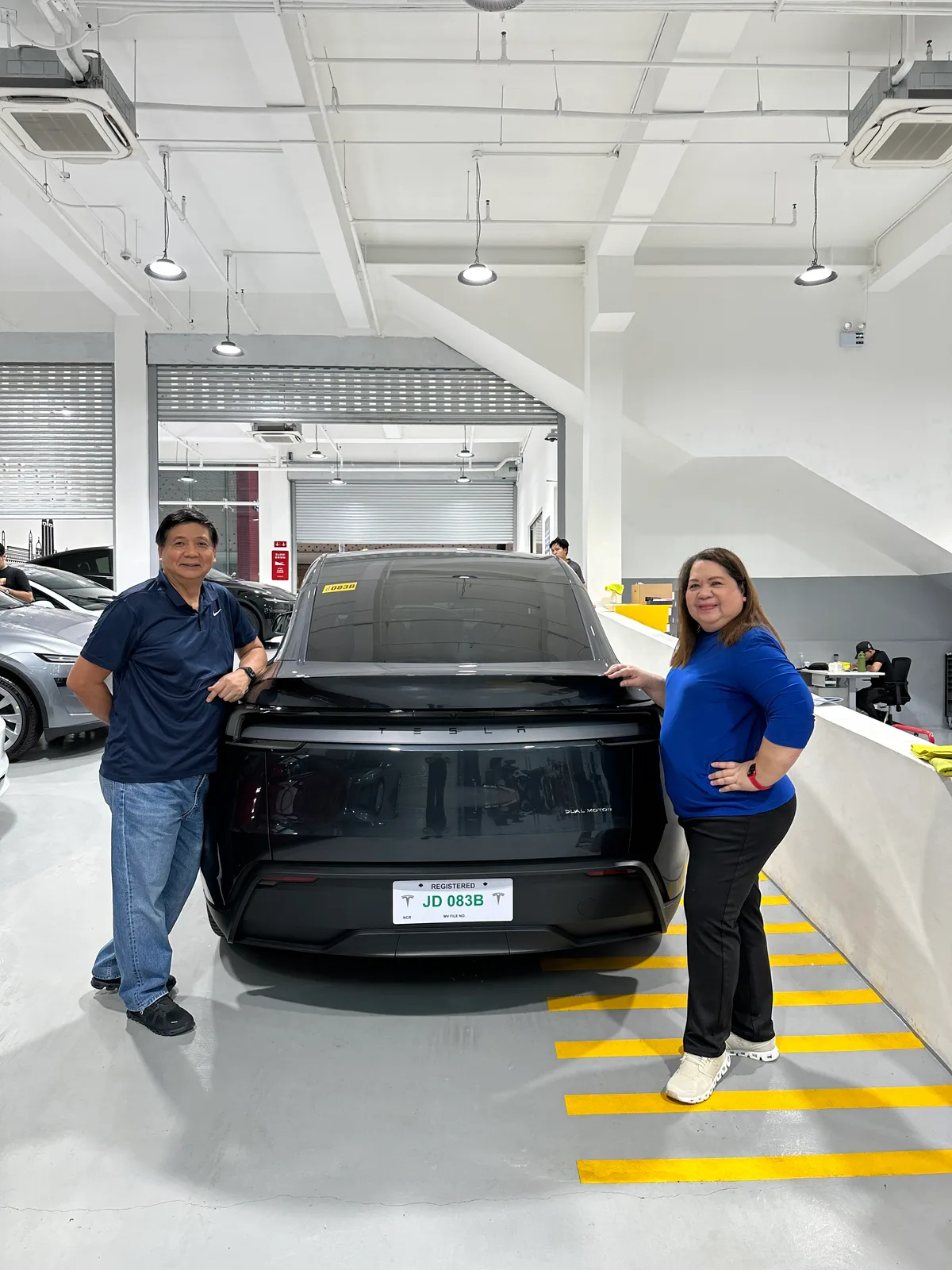 Doc Dennis and his wife posing at the rear of their Stealth Gray Model Y showing the Dual Motor badge