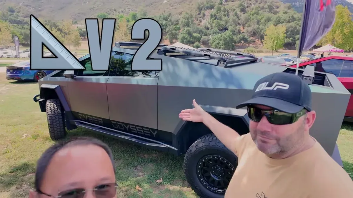 Two men standing in front of the matte gray Cyber Odyssey V2 Cybertruck with roof rack and off-road tires at an outdoor event