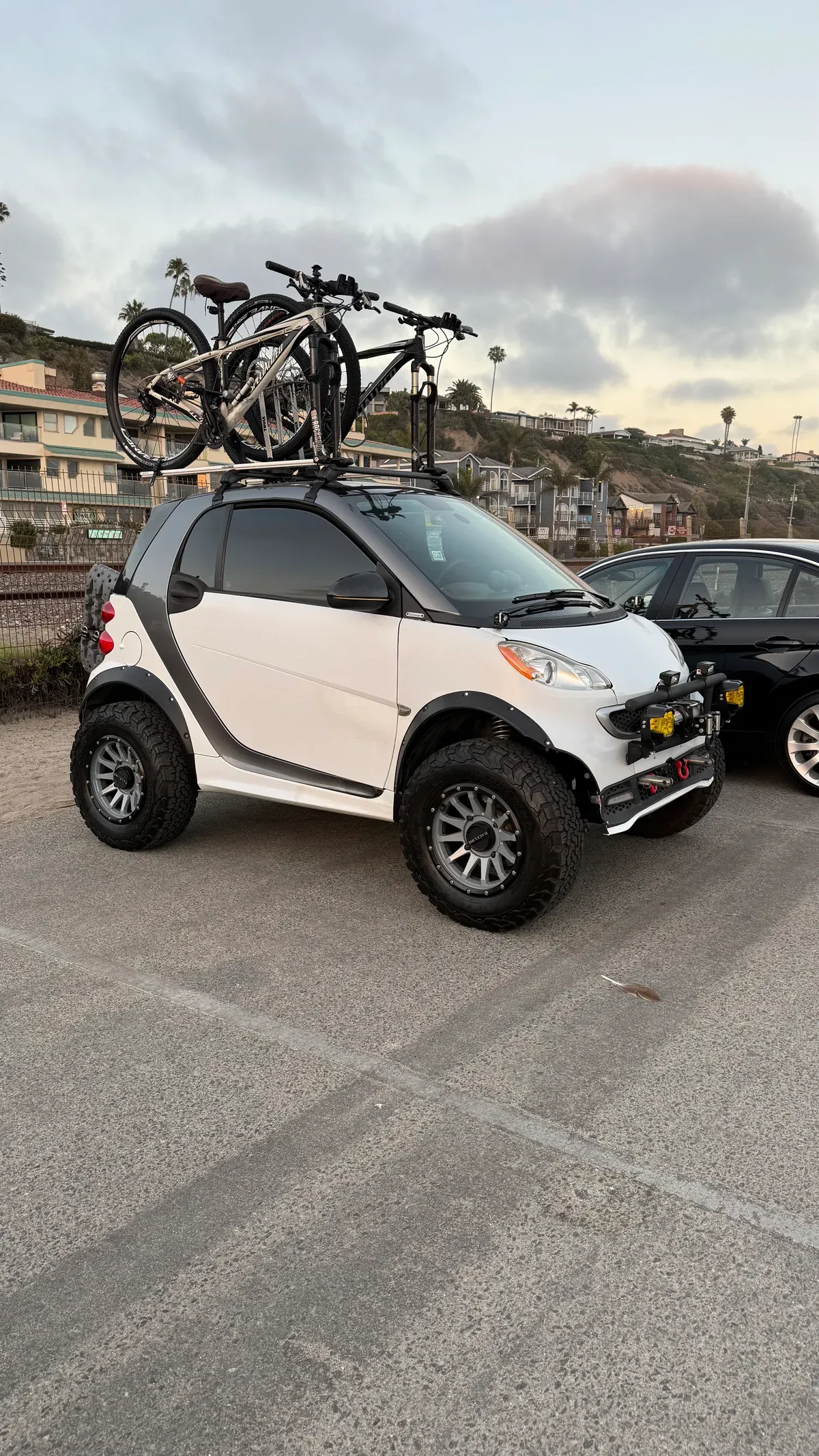 Customized white Smart car with oversized off-road tires and a roof rack carrying two mountain bikes at Dana Point beach parking