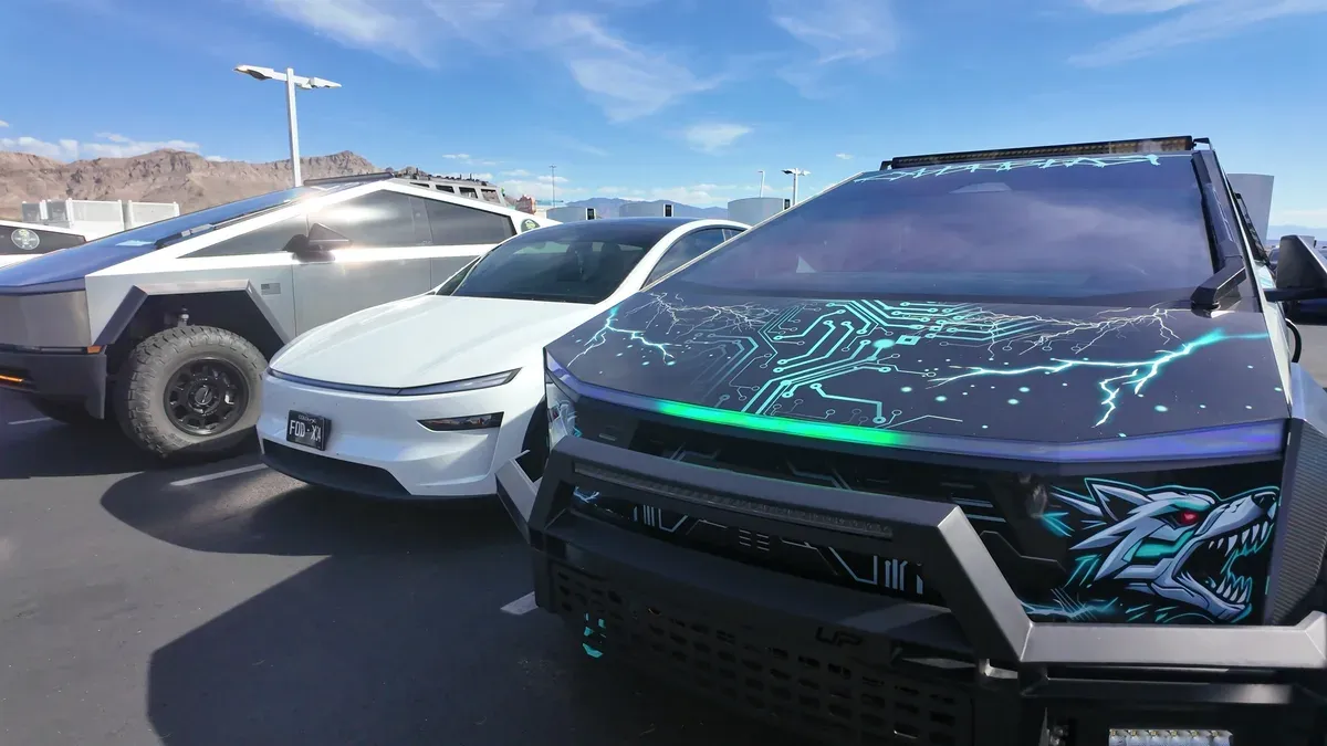 Front of a wrapped Cybertruck with a black Grok AI-designed wrap, teal circuit-board and lightning graphics, a snarling wolf on the bumper and a lightbar, parked beside a stainless Cybertruck and a white Tesla