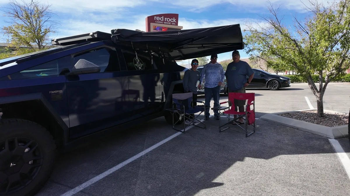 Dark Cybertruck with a side awning deployed at the Red Rock Casino meetup, three owners standing in the shade beside folding camp chairs