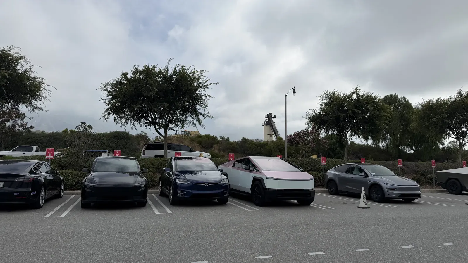 A lineup of Cybertrucks and Teslas, including a pink-wrapped Cybertruck, parked in a lot on a cloudy SoCal day.