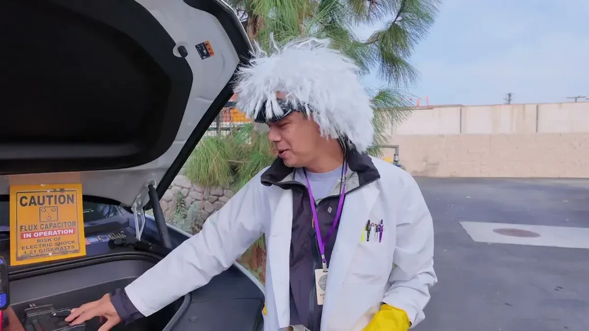 Person dressed as Doc Brown pointing into the open frunk of a white Rivian with a Caution Flux Capacitor sign