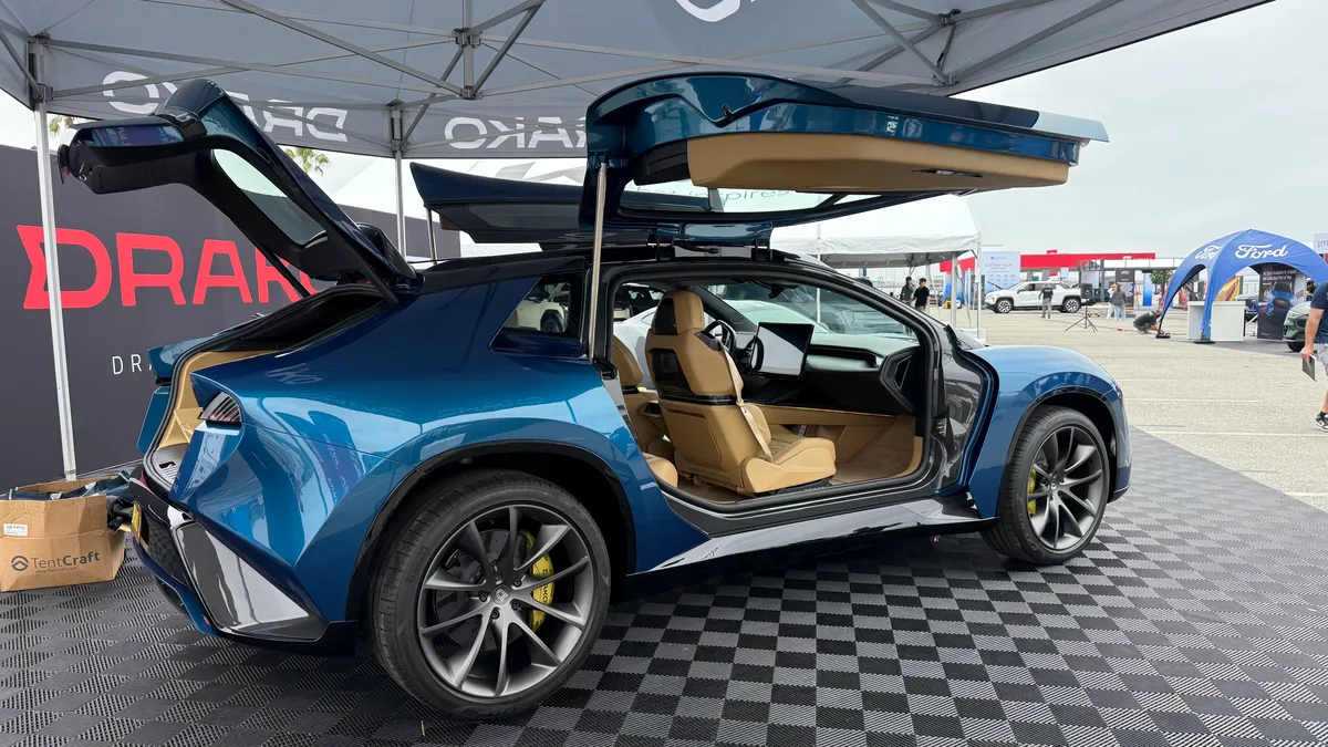 Blue Drako Dragon EV with gull-wing doors and rear hatch open, yellow brake calipers and tan interior on a checkered display floor