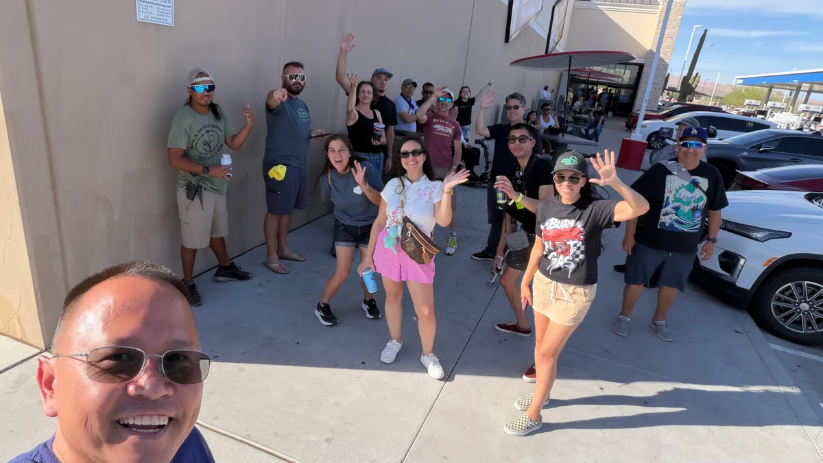 About fifteen EV owners waving at the camera for a group selfie beside a wall at a roadside Chevron stop on the Vegas run