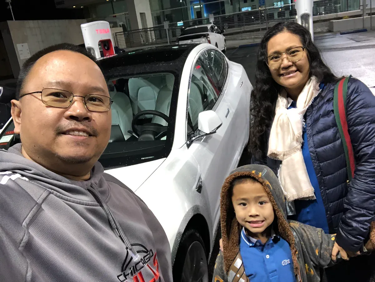 Sherwin, Abby, and their son together in a selfie next to the white Model 3 at the Tesla dealership