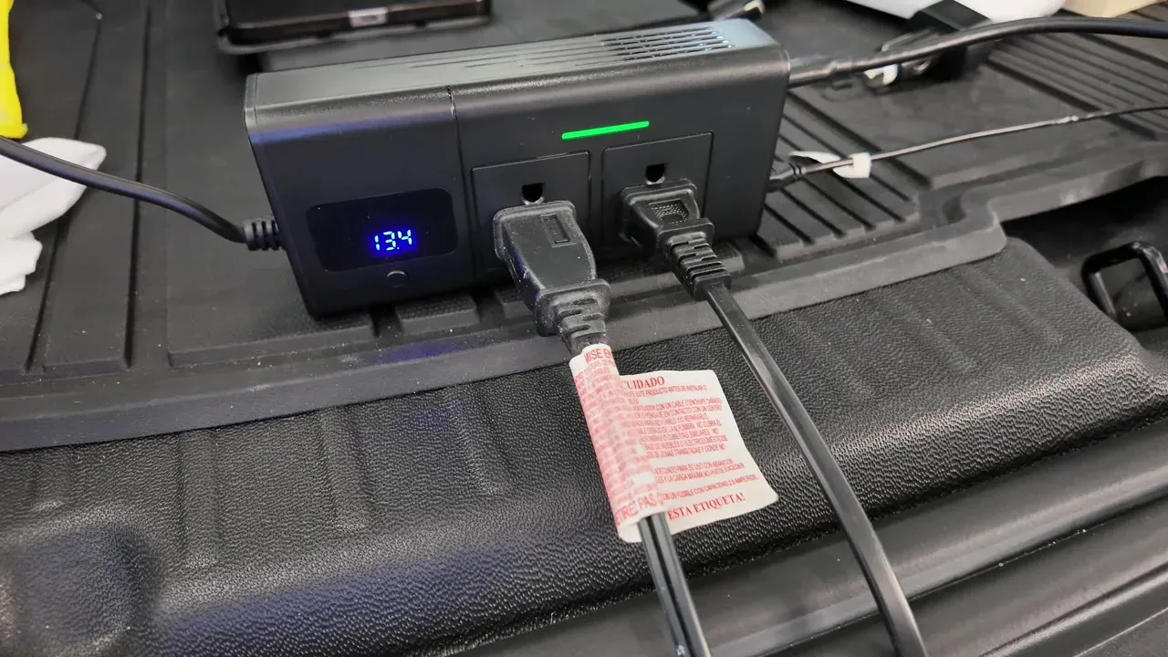 The Hansshow 200W inverter sitting in the trunk of a Model Y with both 110V AC outlets occupied, its green power light on and the voltage display reading 13.4.