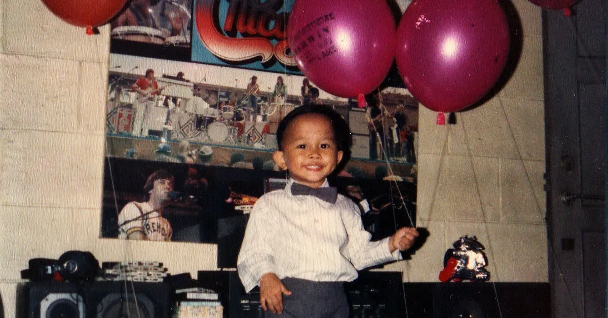 Happy young boy in a bowtie holds balloons in front of a band poster and speakers.