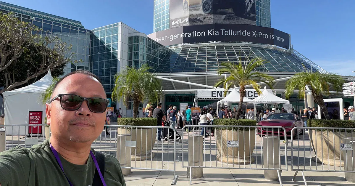 Man in sunglasses takes selfie at outdoor auto show entrance with convention center, Kia banner.