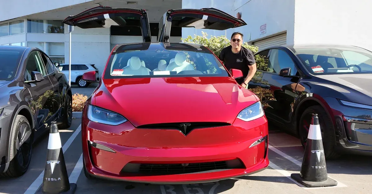 A man stands beside a red Tesla Model X with its falcon wing doors open in a parking lot.