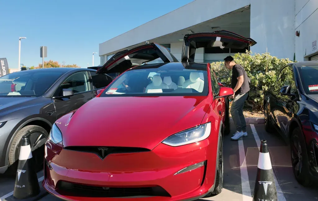 Red Tesla Model X with falcon wing doors open, parked outdoors; man looks inside.