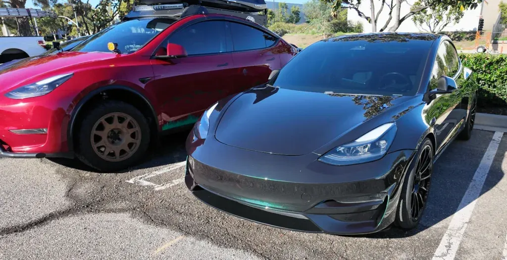 A red off-road Tesla Model Y with a roof rack parked next to a dark sporty Tesla Model 3.