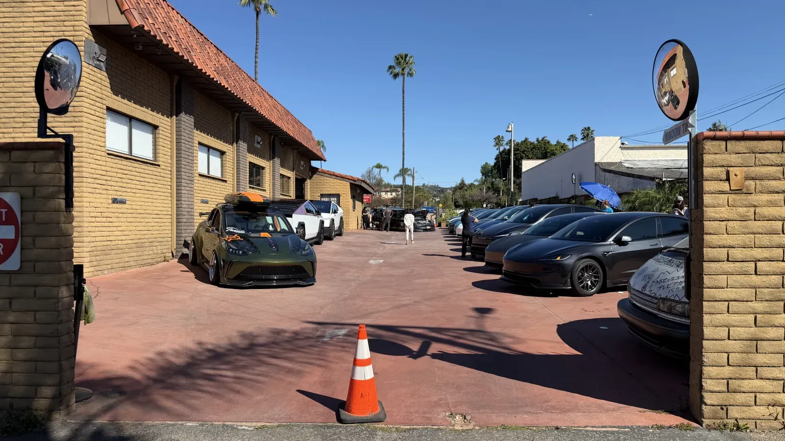 Wide view of the Cars and Coffee lot with a green Tesla Model 3 in the foreground, an orange traffic cone, and a row of Model Ys and Model 3s under a blue umbrella canopy.
