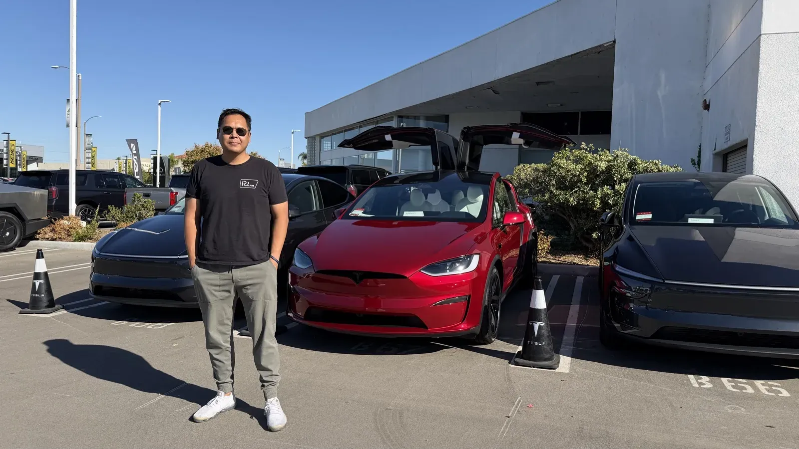 Ultra Red 2026 Tesla Model X Plaid with falcon wing doors open at the Tesla delivery lot, a man standing in front of it, a Cybertruck and other Teslas parked nearby.