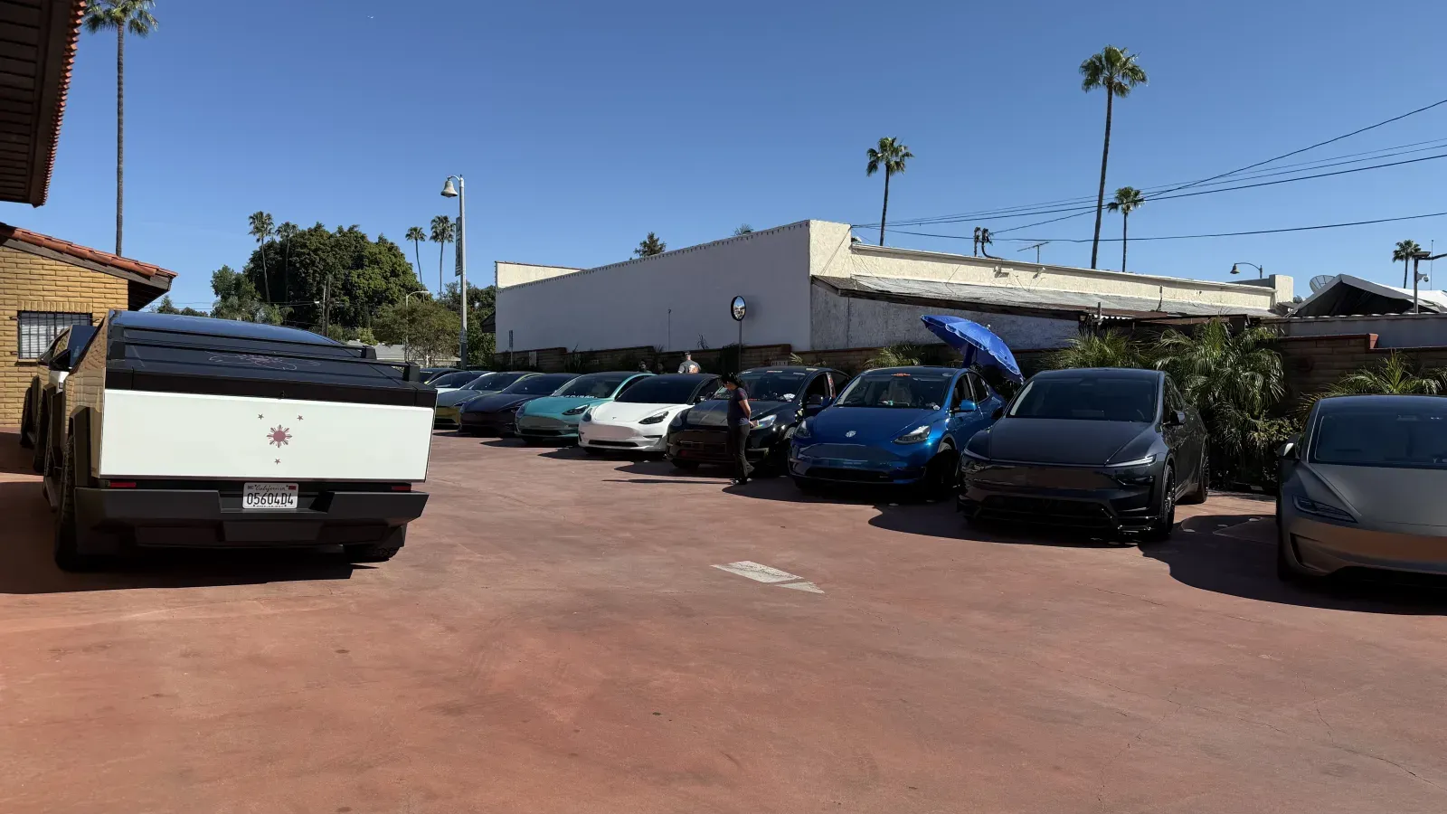 Wide angle of the lot showing our Tesla Cybertruck with a Filipino sun decal on the tailgate in the foreground, with a row of teal, white, and blue Teslas behind under a clear sky.