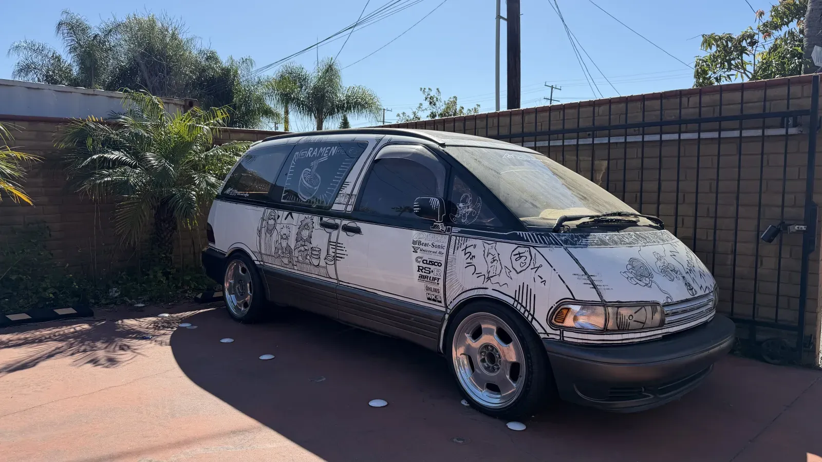White Toyota Previa with hand-drawn anime and manga line art covering the entire body, parked against a wooden fence with deep-dish wheels and a lowered stance.