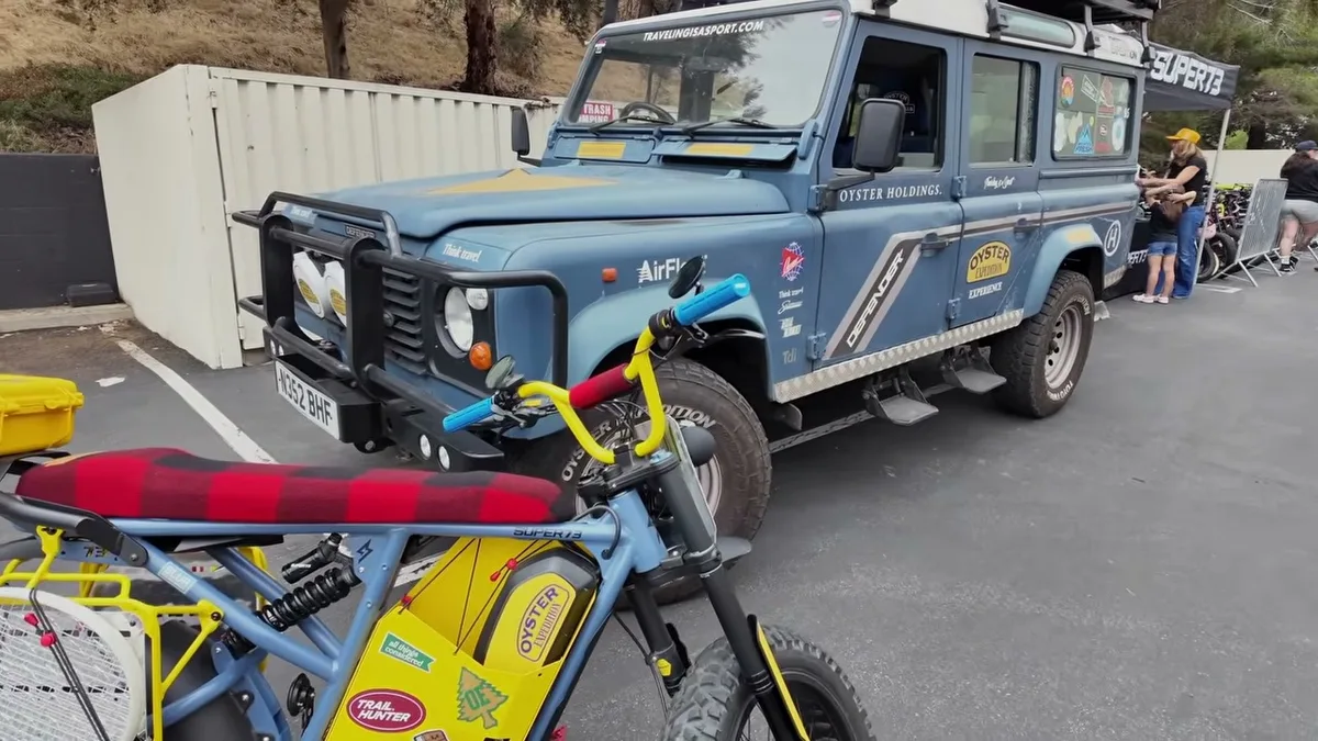 Blue 1988 Land Rover Defender 110 Oyster Expedition with roof rack, next to a blue and yellow Super73 electric bike.