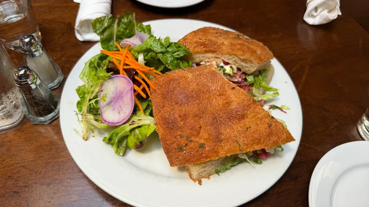 Sandwich on focaccia with a side salad on a white plate at a lunch table