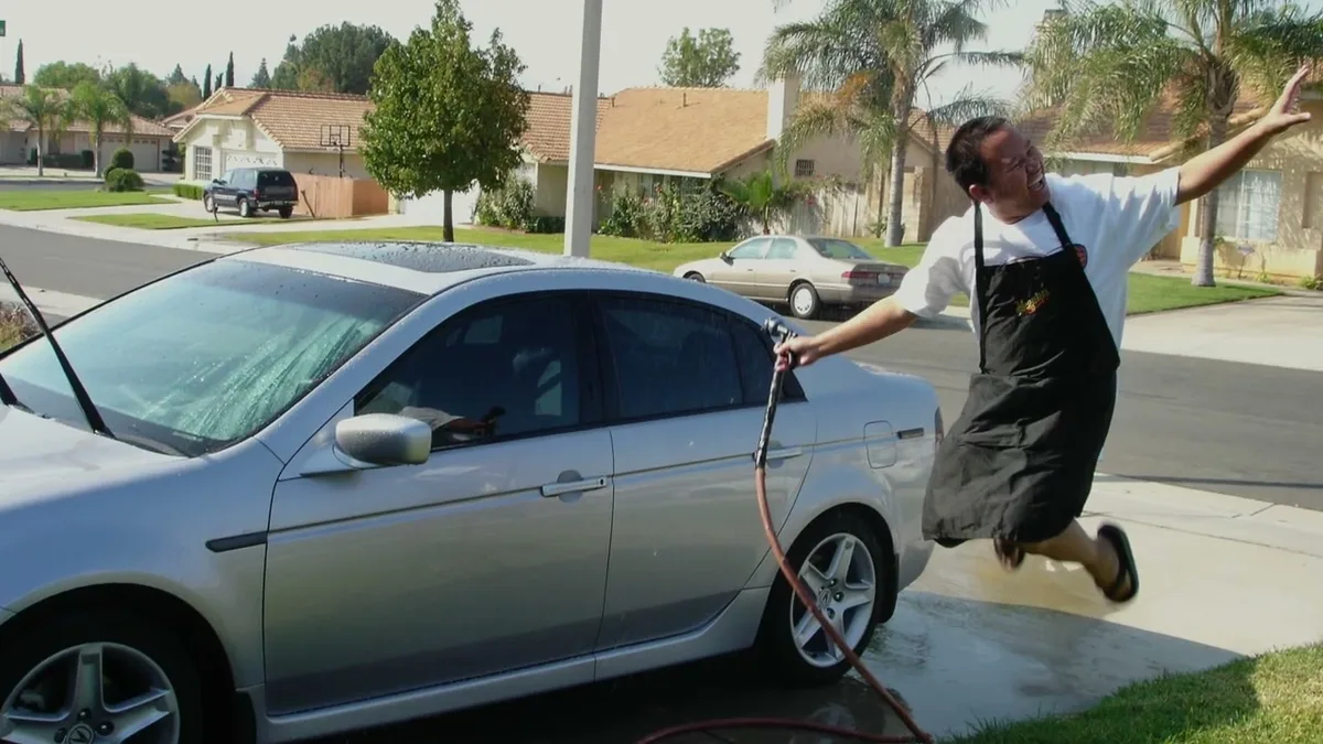 Sherwin in an apron jumping and spraying water on a silver sedan in a suburban driveway