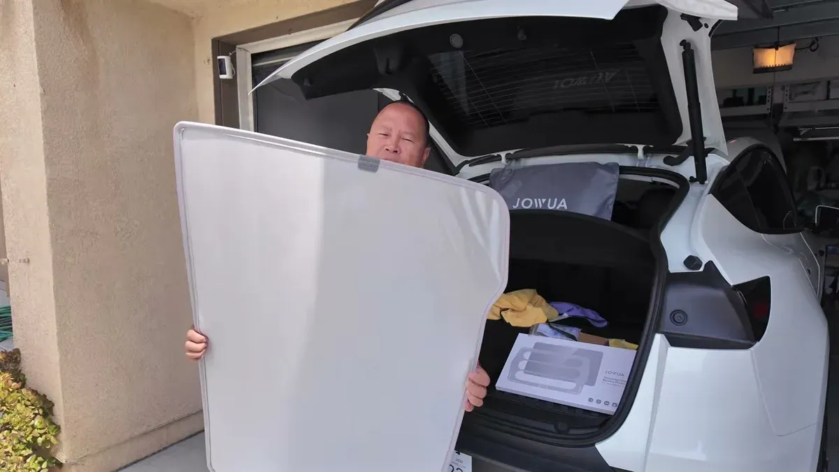 Man holding a large white sunshade in front of a white Tesla Model Y's open trunk with accessories