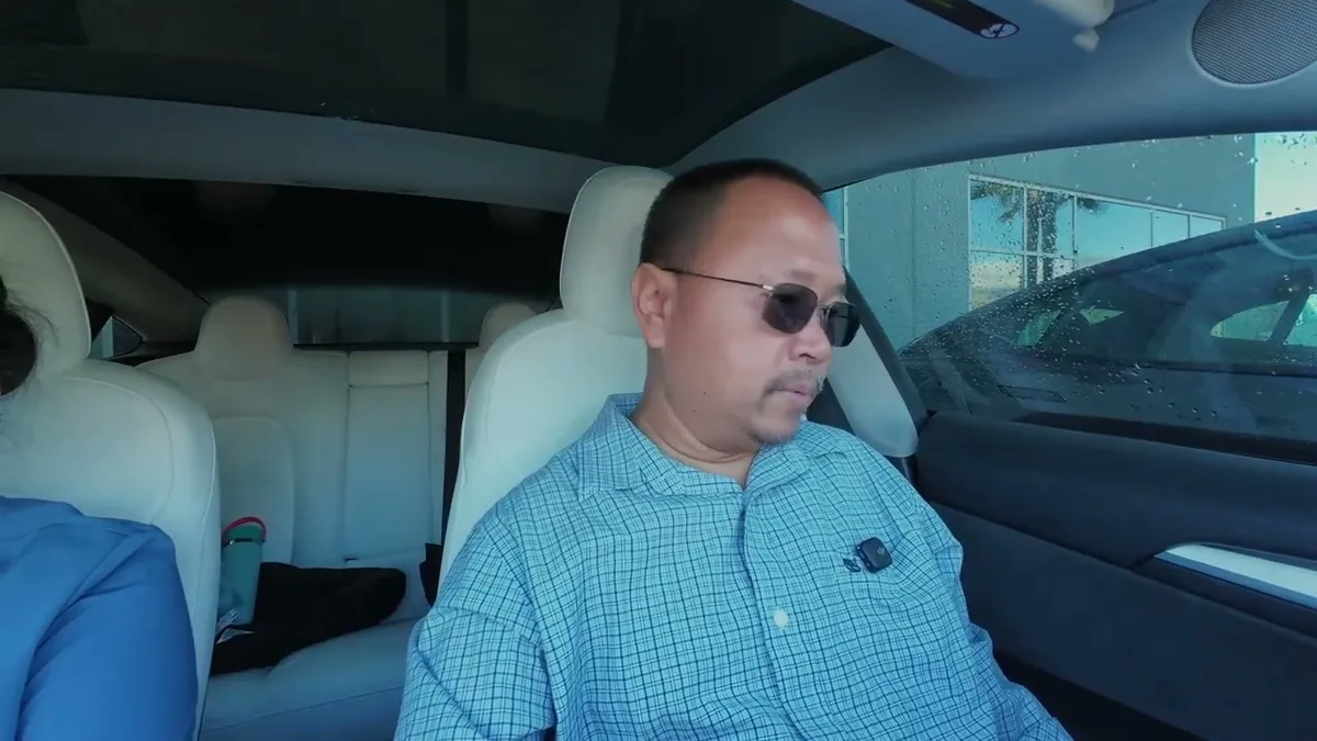 Man in dark sunglasses driving lemon law Model 3 loaner, wet exterior visible outside