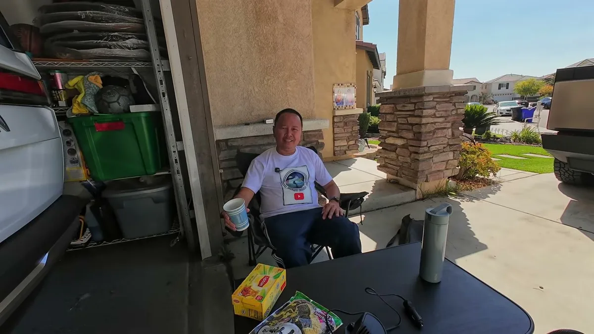 Man relaxing in black camping chair outside garage holding a blue cup, white Tesla Model Y in background