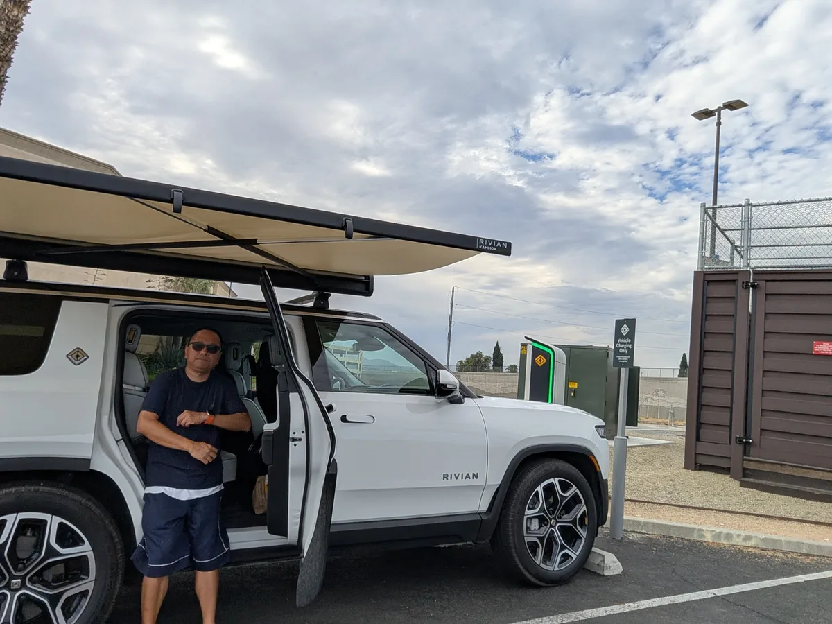 Sherwin standing next to the white Rivian R1S with awning extended at an EV charging station