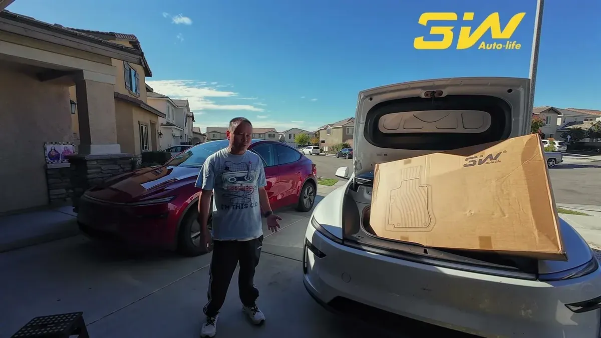 Man gesturing toward the 3W floor mat box in the open frunk of a white Tesla Model Y, red Model Y parked behind