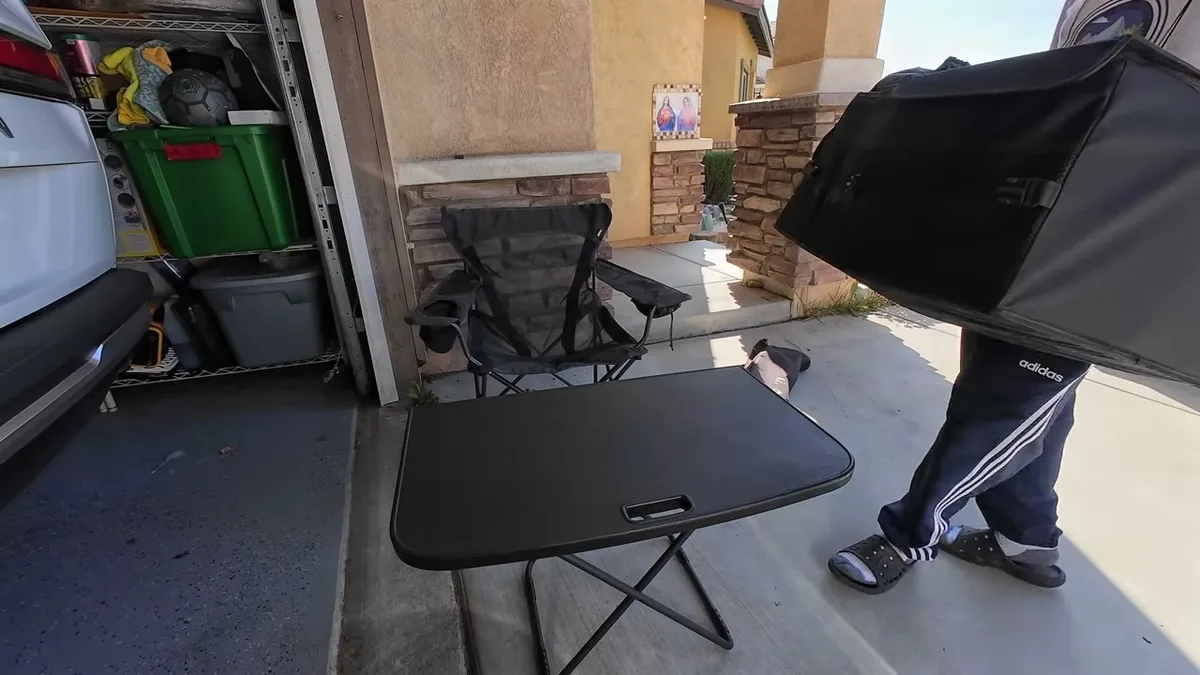 Man unpacking camping gear next to foldable table and camping chair outside garage