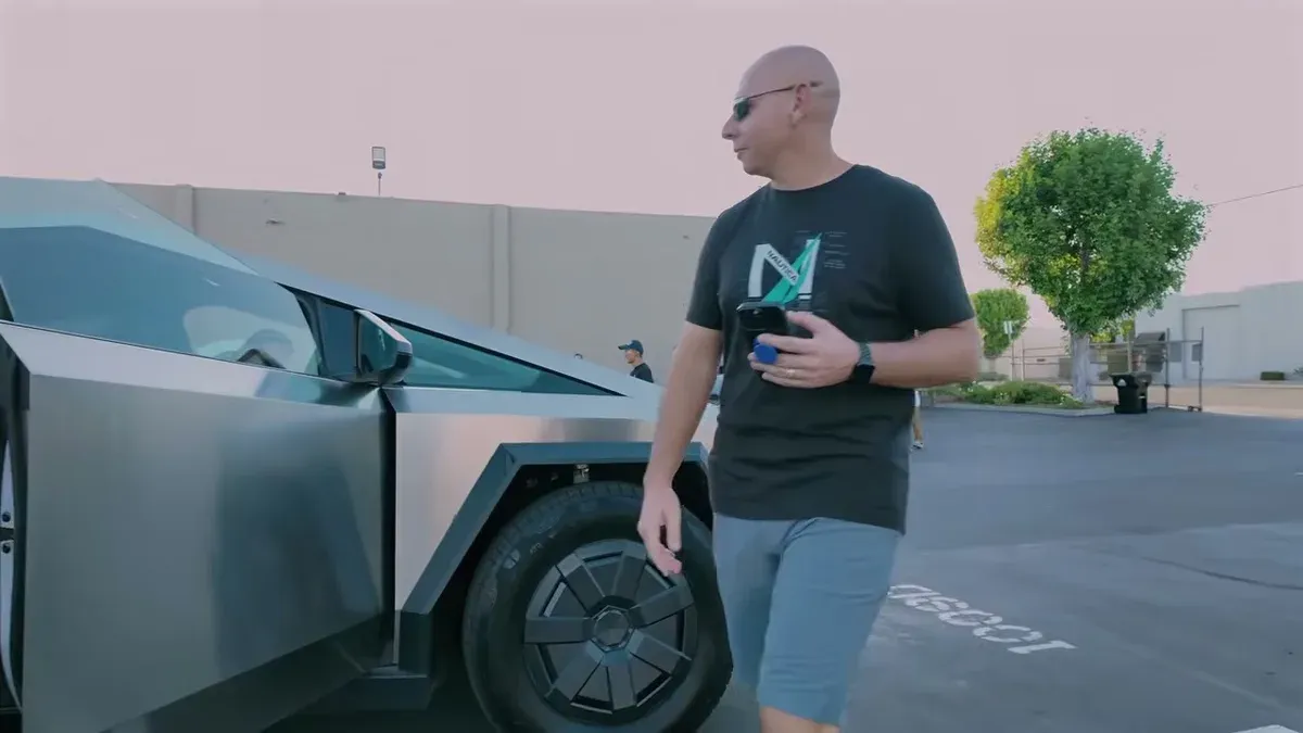 Man walking past silver Tesla Cybertruck in sunny outdoor parking lot with trees.