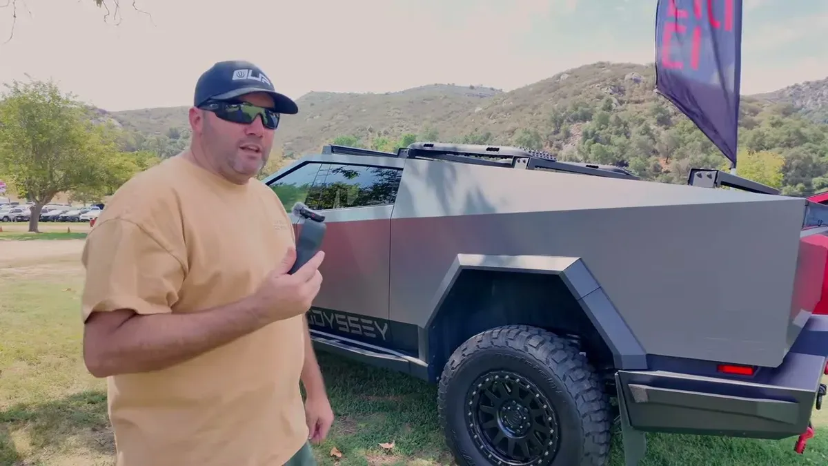 Brian standing next to the Cyber Odyssey V2 Cybertruck in side profile, sunglasses and tan shirt, roof rack and off-road build visible