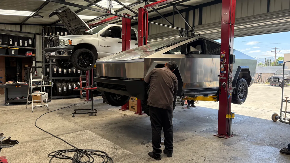 Mechanic working on a lifted Tesla Cybertruck in a garage with another truck on a lift.
