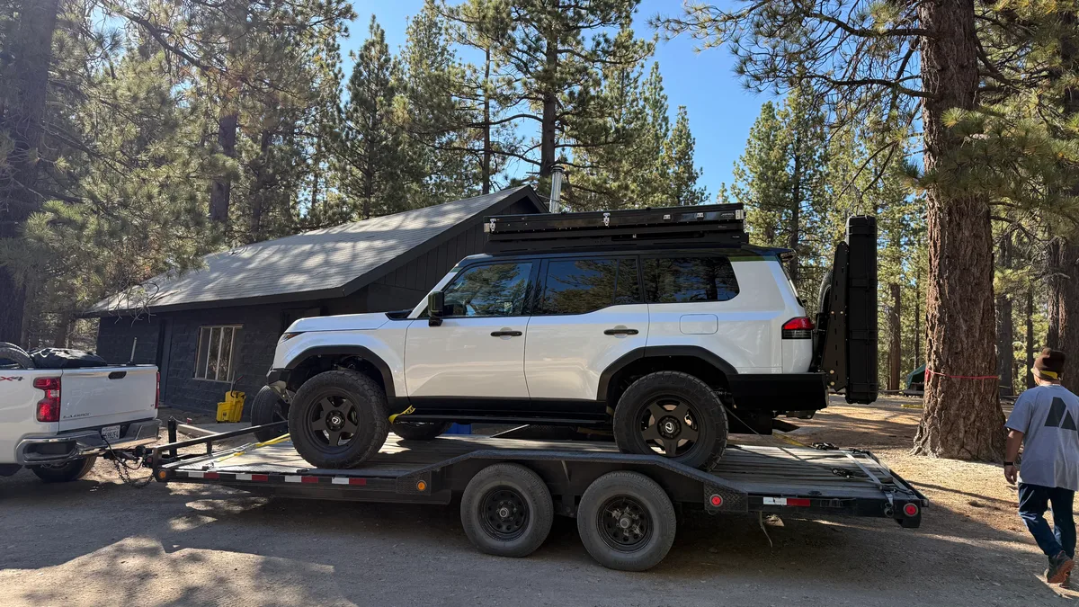 Modified white Ford Bronco loaded on a flatbed trailer next to a forest cabin