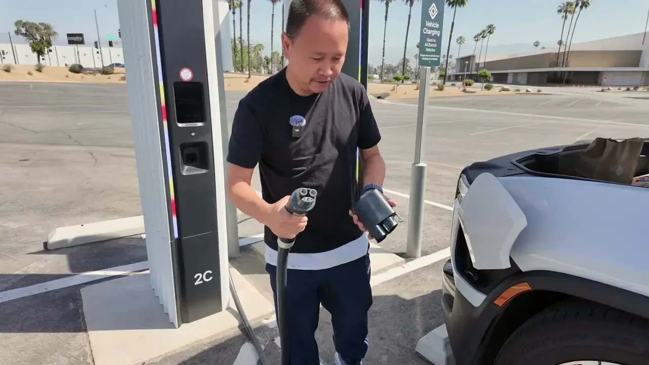 Sherwin holding the Rivian CCS-to-NACS adapter at the San Bernardino Rivian Adventure Network, with the Gen 2 R1S charge port flap open behind him.