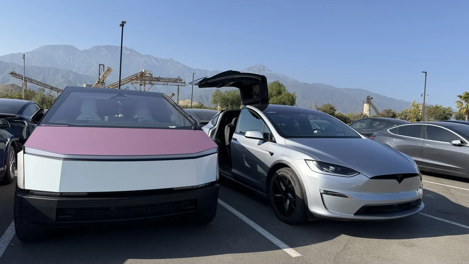 A pink-wrapped Cybertruck parked beside the silver Tesla Model X loaner with a falcon wing door open, mountains behind.