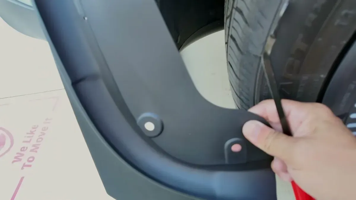 Hand holding black mud flap with pre-drilled holes against a white car.