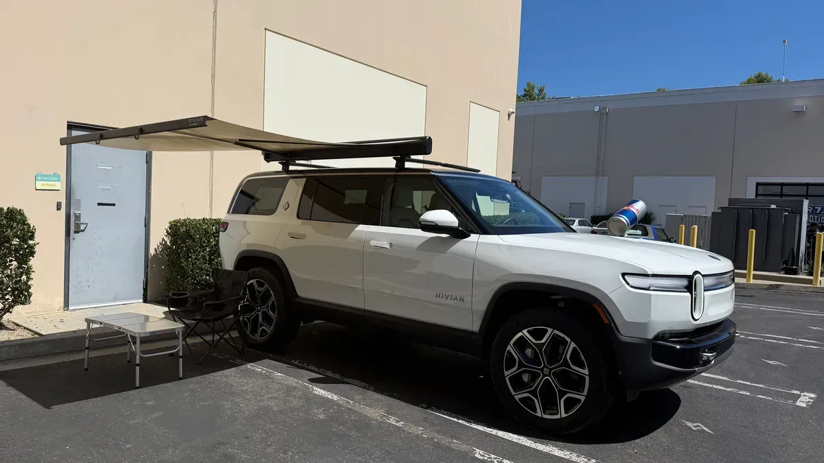 White Rivian R1S with side awning fully extended over camp table and chair in sunny parking lot