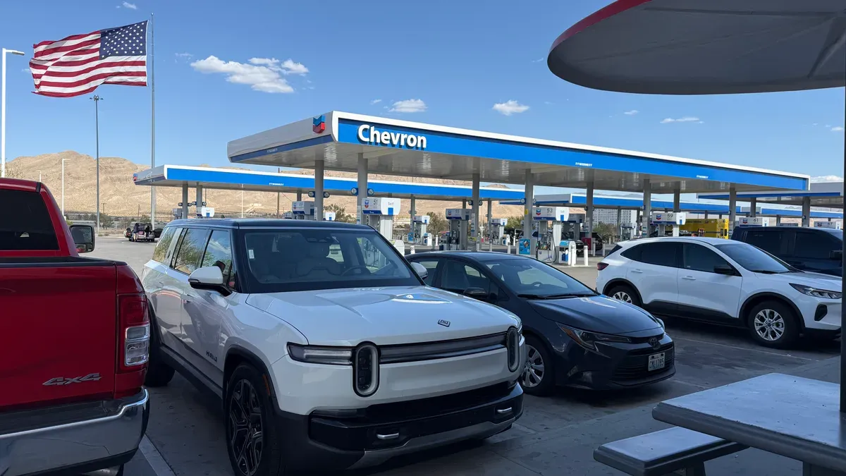 White Rivian R1S parked at a Chevron gas station with a large American flag in the background