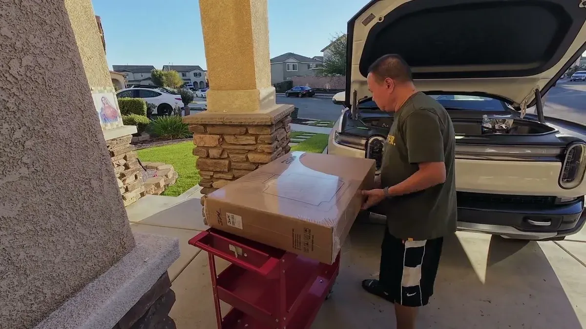 Man lifting 3W floor mat box from red cart next to open-frunk white Rivian R1S in driveway