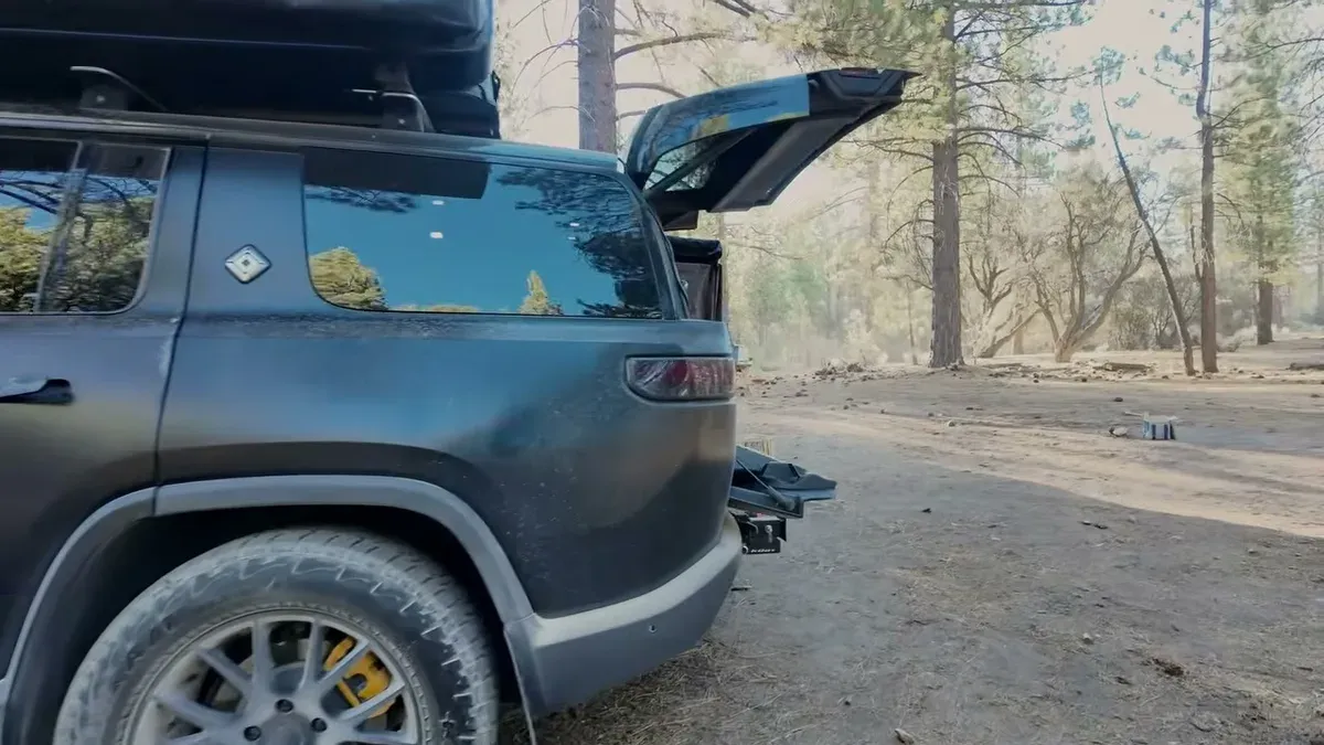 Matte black Rivian R1S with open tailgate and roof rack parked in a sun-dappled pine forest