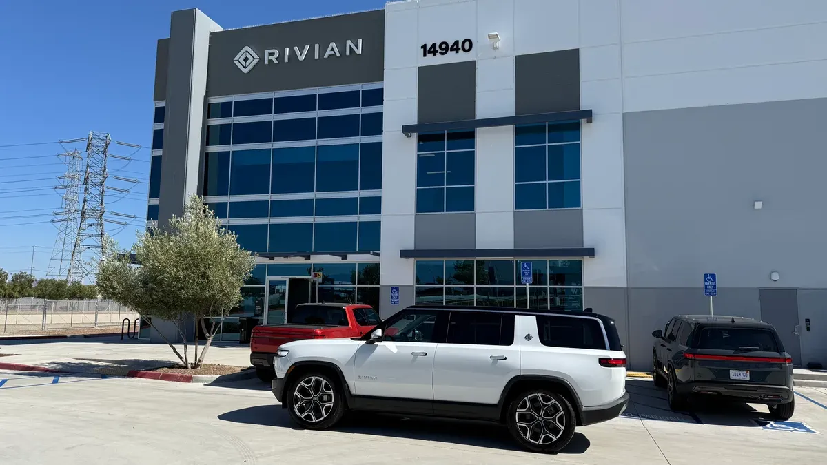 White Rivian R1S parked outside a Rivian service center, with a red R1T and a dark grey R1S in the lot