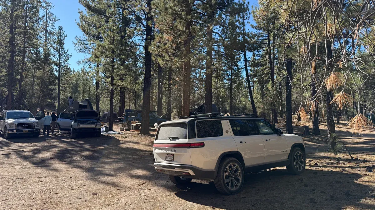 White Rivian R1S rear view with other Rivians and campers packing up in the pine forest background