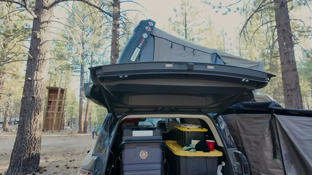 View from inside Rivian R1S cargo area looking out at deployed rooftop tent and pine forest campsite