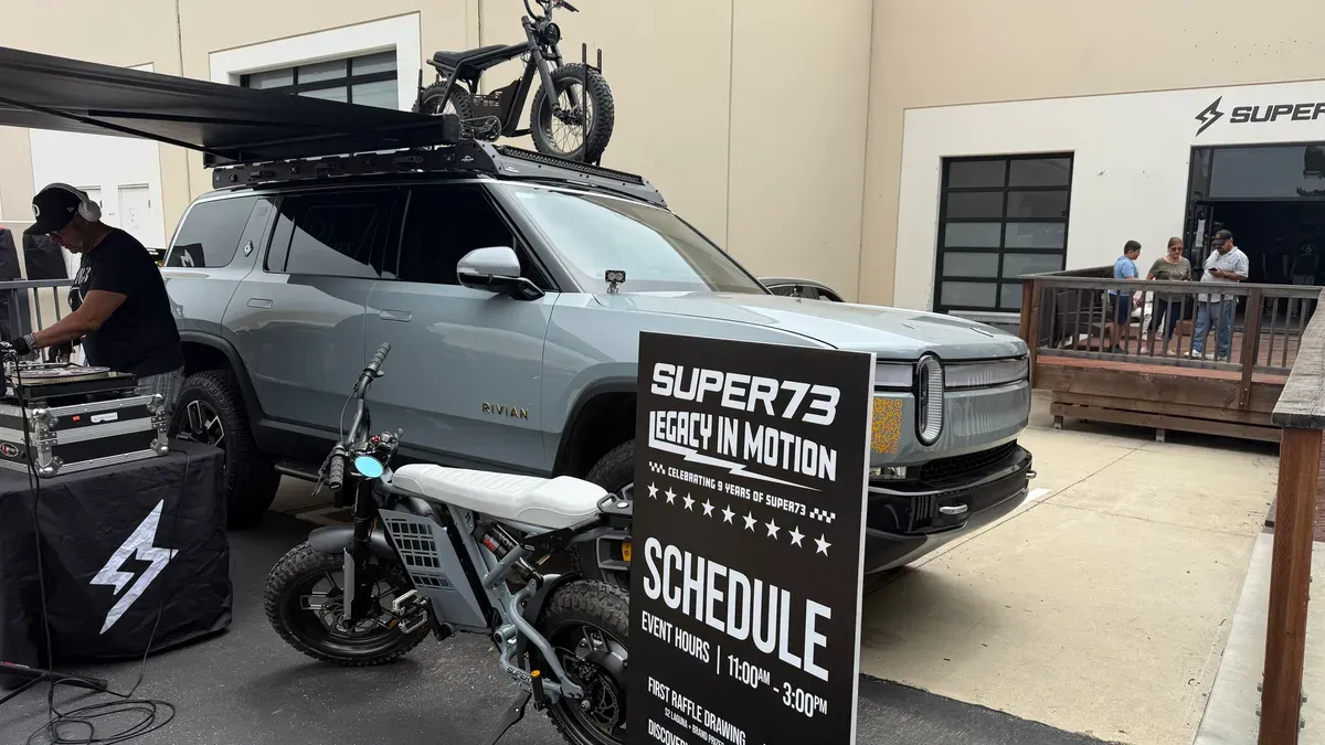 Rivian R1S SUV with Super73 electric bikes on roof and in front at an outdoor event with a DJ and schedule sign.