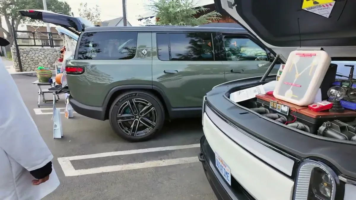 Two Rivian SUVs parked side by side with trunks open, white R1T frunk showing a Flux Capacitor prop, dark green R1S tailgate open